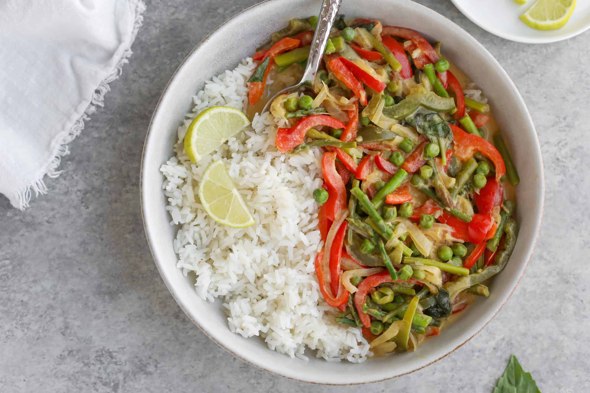 Spring Vegetable Thai Green Curry 20 A Bowl Of White Rice And Thai Green Curry Featuring Red Peppers, Peas, And Green Beans. Garnished With Lime Slices, A Fork Rests In The Bowl On A Gray Textured Surface. A White Cloth And Extra Lime Slices Add To The Scene In The Background.
