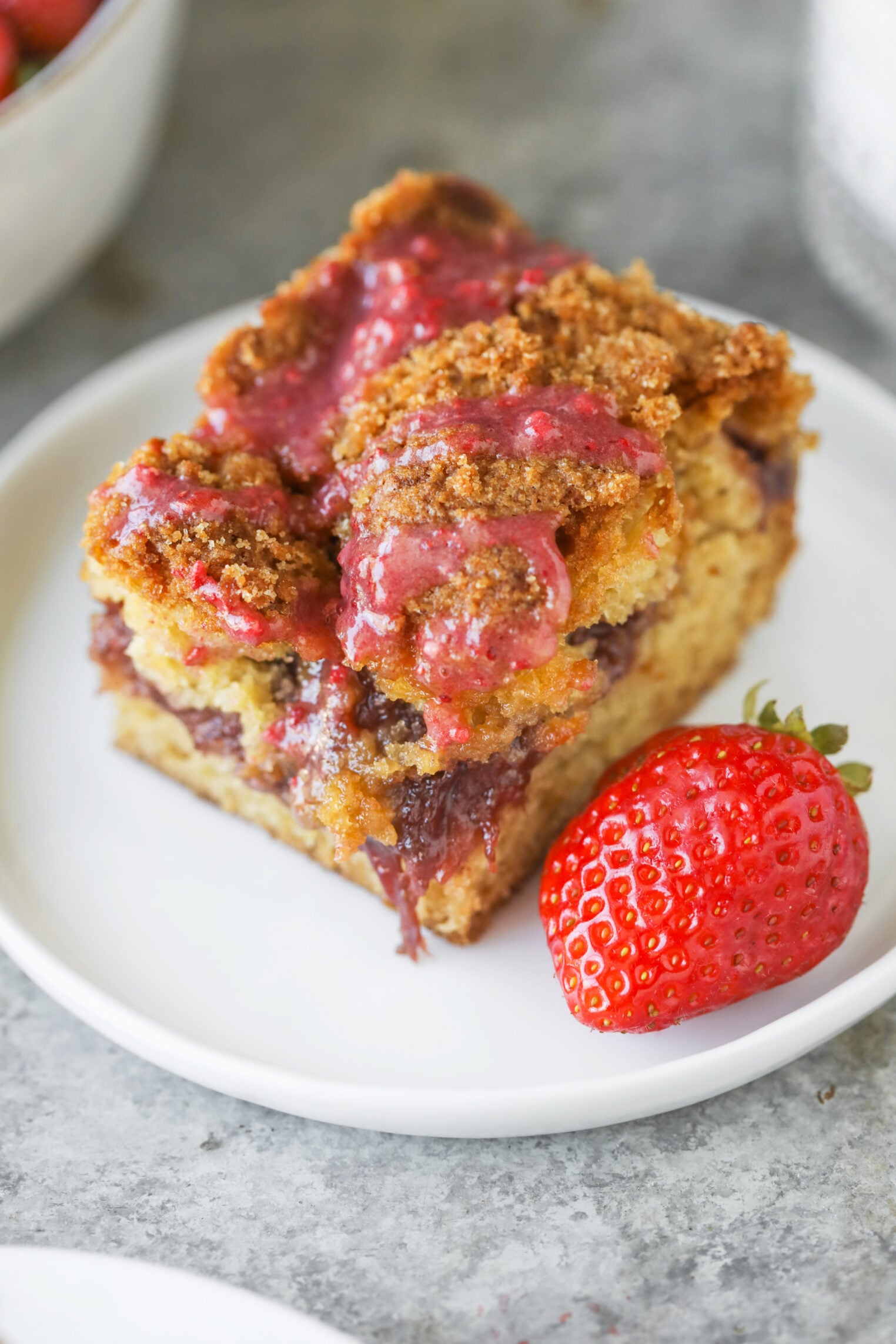 A Slice Of Crumbly Strawberry Coffee Cake With A Layer Of Berry Filling And Pink Glaze, Served On A White Plate With A Fresh Strawberry Beside It.