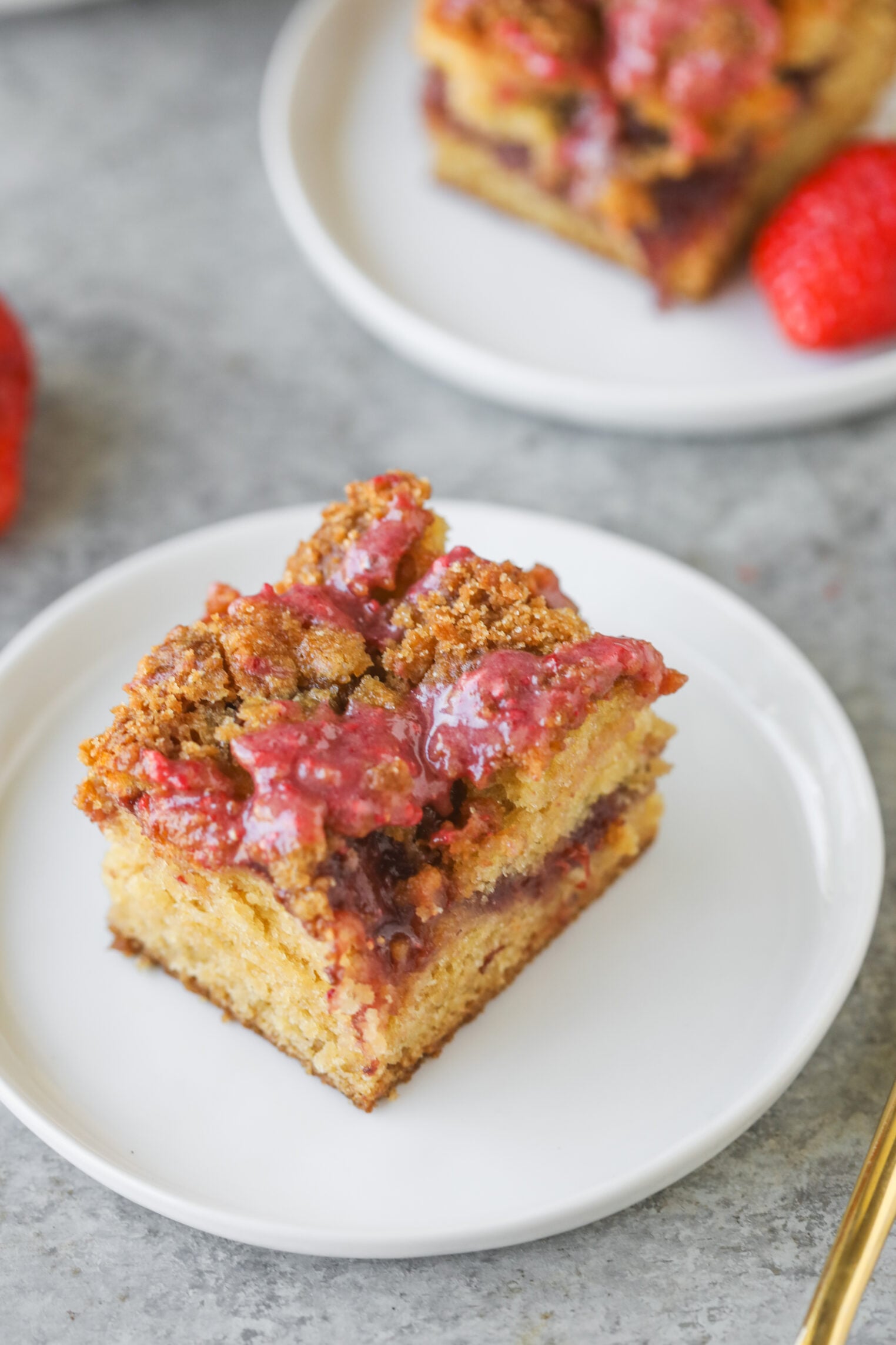 A Square Slice Of Strawberry Coffee Cake With A Crumb Topping And A Layer Of Strawberry Filling Sits On A White Plate. The Moist Cake Features A Golden-Brown Crumb And Is Finished With A Drizzle Of Strawberry Sauce On Top.
