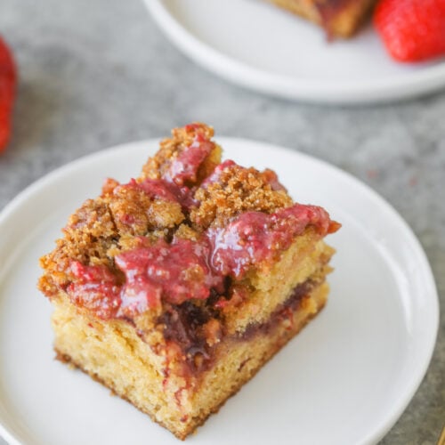 A Square Slice Of Strawberry Coffee Cake With A Crumb Topping And A Layer Of Strawberry Filling Sits On A White Plate. The Moist Cake Features A Golden-Brown Crumb And Is Finished With A Drizzle Of Strawberry Sauce On Top.