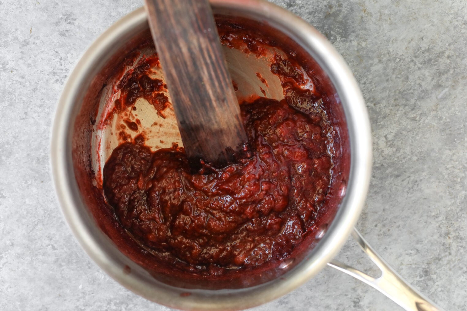 A Stainless Steel Saucepan Containing A Thick, Red Strawberry Coffee Cake Sauce Being Stirred With A Wooden Spatula, Set On A Gray Textured Surface.