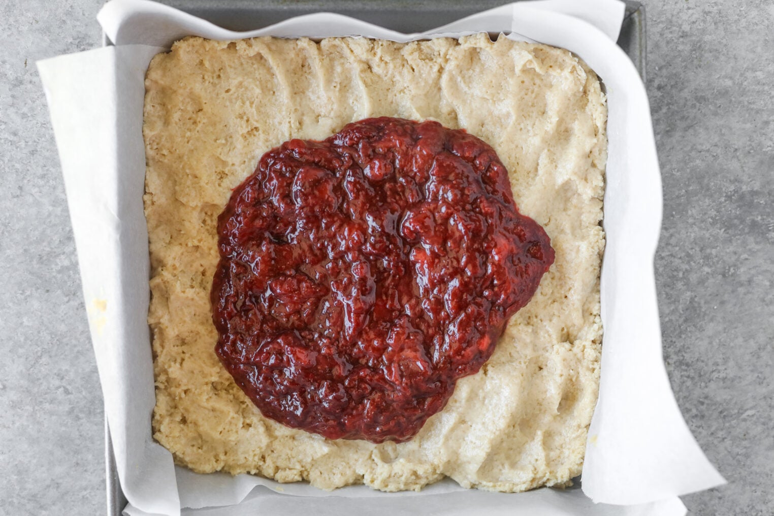 A Square Baking Pan Lined With Parchment Paper Holds A Pressed, Unbaked Oat Crust Topped With A Generous Dollop Of Chunky Strawberry Coffee Cake Jam, Ready To Be Spread.