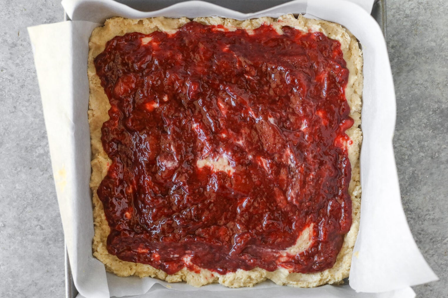 A Square Baking Pan Lined With Parchment Paper, Filled With A Layer Of Crust Topped With A Generous Spread Of Red Fruit Jam, Reminiscent Of A Strawberry Coffee Cake.