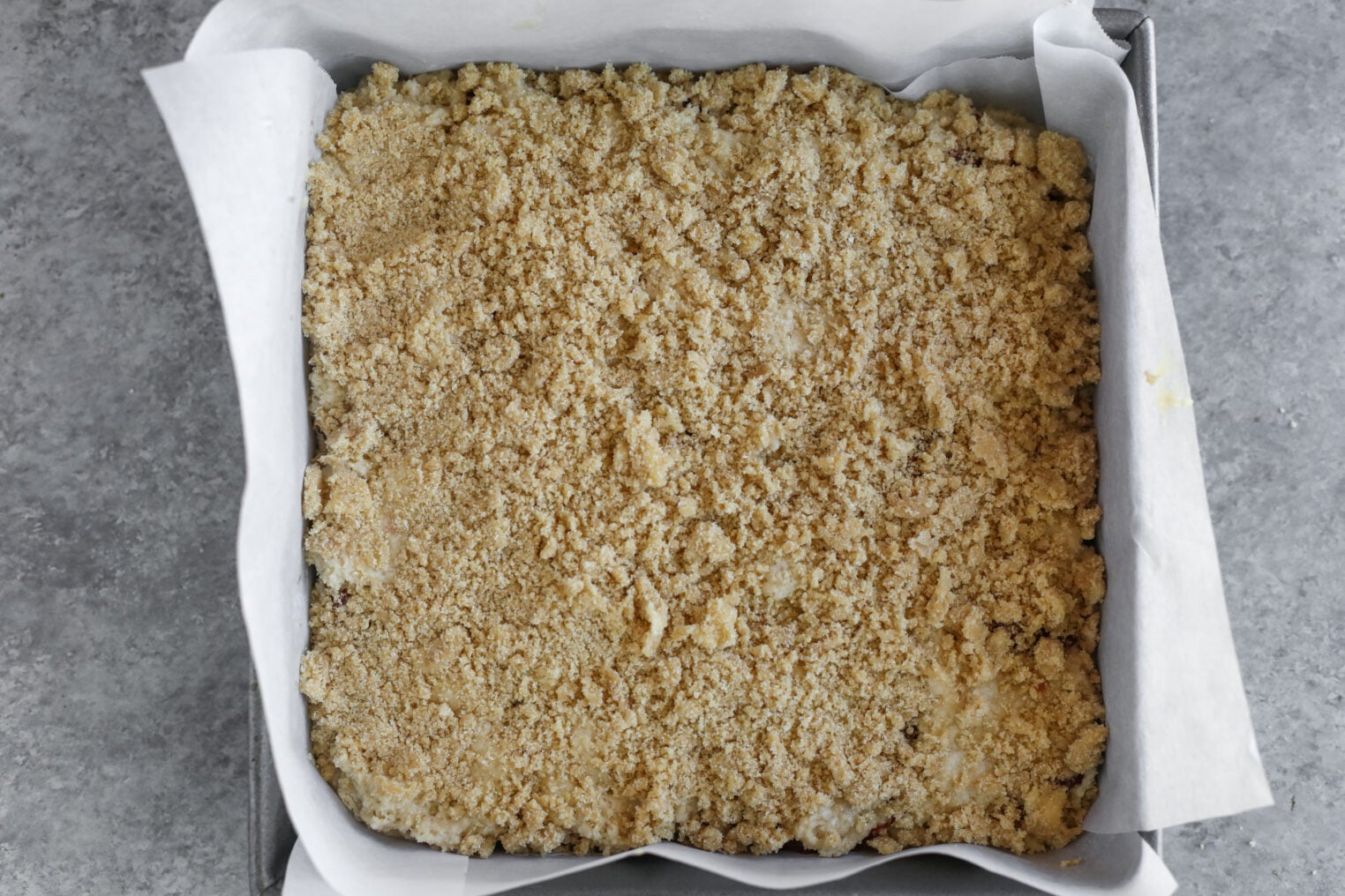 A Parchment Paper-Lined Square Baking Pan Filled With Unbaked Crumbly Streusel Topping Sits Atop A Gray Countertop, Ready To Become A Delicious Strawberry Coffee Cake.