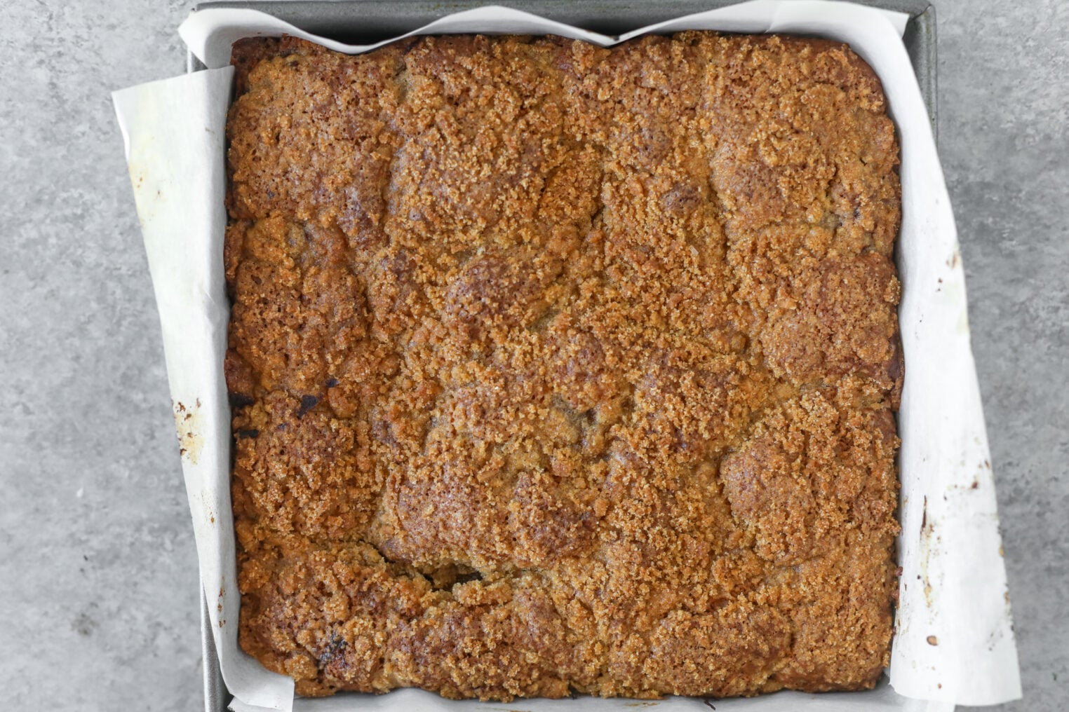 A Square, Golden-Brown Strawberry Coffee Cake With A Crumbly Topping Is Shown In A Parchment-Lined Baking Pan On A Gray Surface.