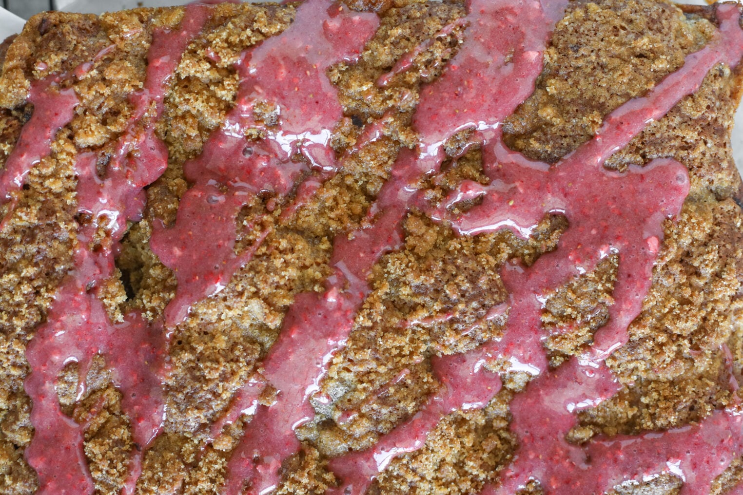 A Close-Up Of A Golden-Brown Strawberry Coffee Cake Topped With A Bright Pink, Glossy Glaze Drizzled In Zigzag Patterns Across The Surface.