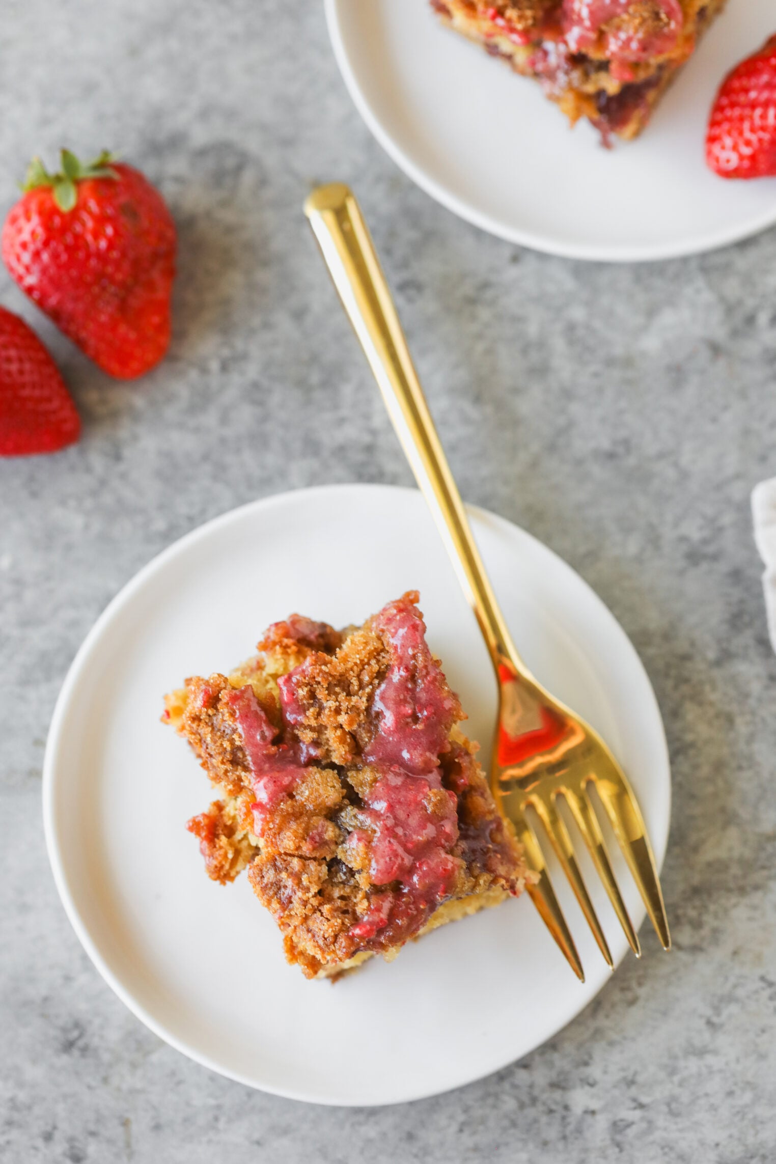 A Square Slice Of Strawberry Coffee Cake Crumble Dessert On A White Plate With A Gold Fork, Surrounded By Fresh Strawberries, On A Gray Textured Surface.