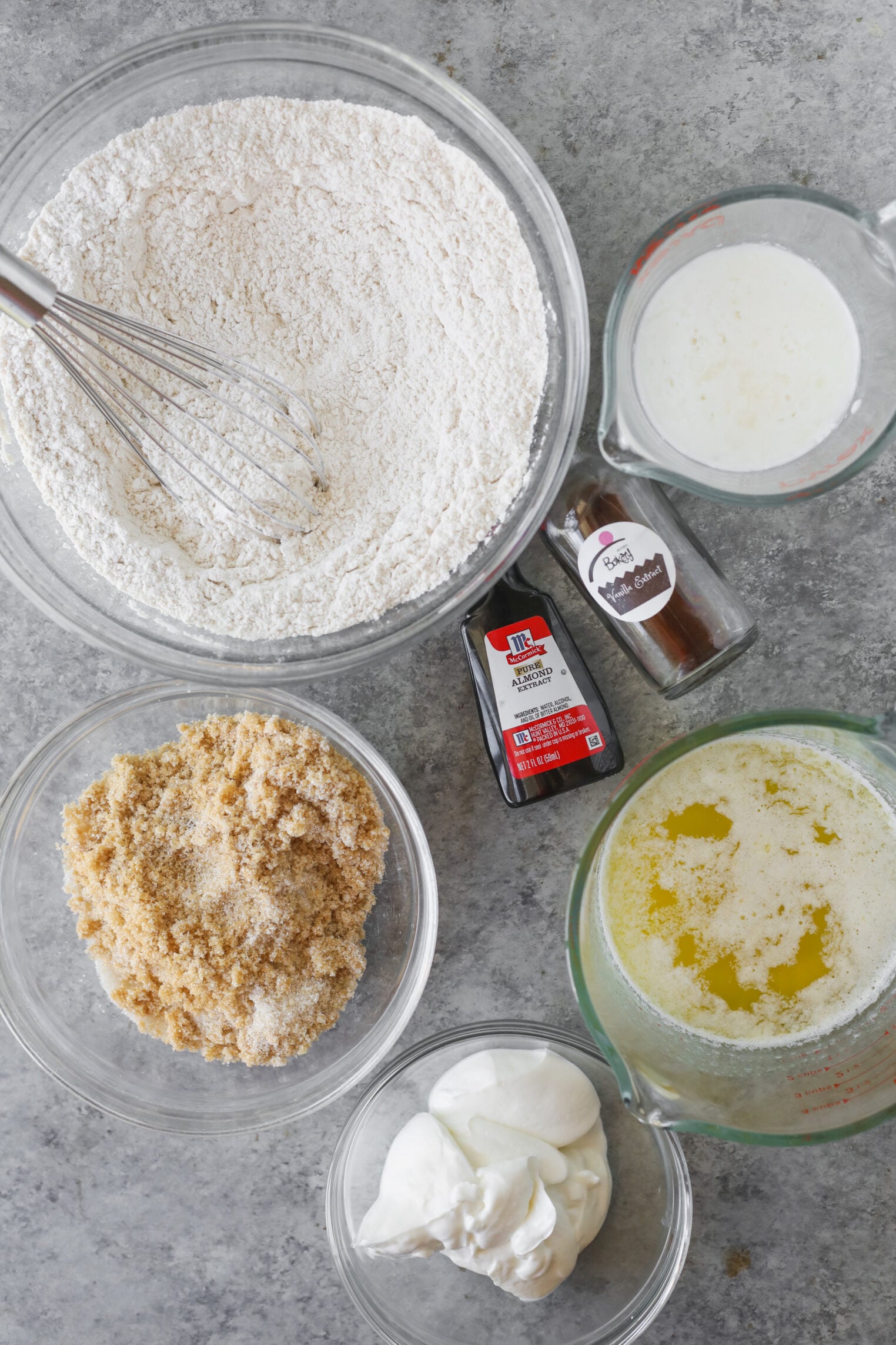 Bowls Of Flour, Brown Sugar, Melted Butter, And Sour Cream—Perfect For Making Strawberry Coffee Cake—Plus A Measuring Cup Of Milk, Vanilla Extract, And Ground Cinnamon Are Arranged On A Gray Countertop.