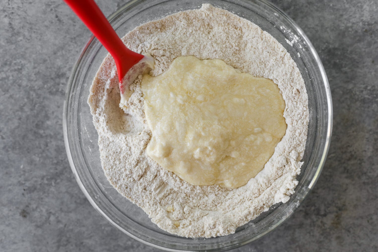 A Glass Bowl Containing Flour With A Well Of Liquid Ingredients In The Center, Ready To Become A Strawberry Coffee Cake. A Red Spatula Rests On The Edge, Poised To Mix Everything Together. The Bowl Sits On A Gray Countertop.