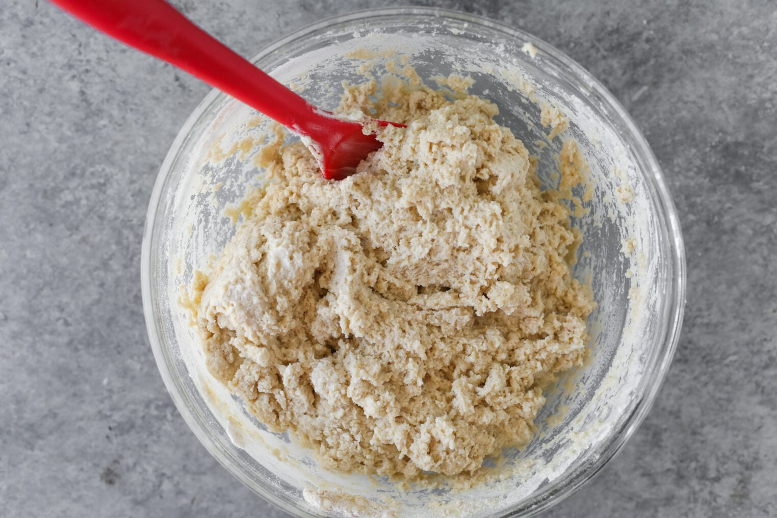 A Glass Bowl Filled With Thick, Pale Strawberry Coffee Cake Dough Being Mixed With A Red Silicone Spatula, Set On A Gray Countertop.