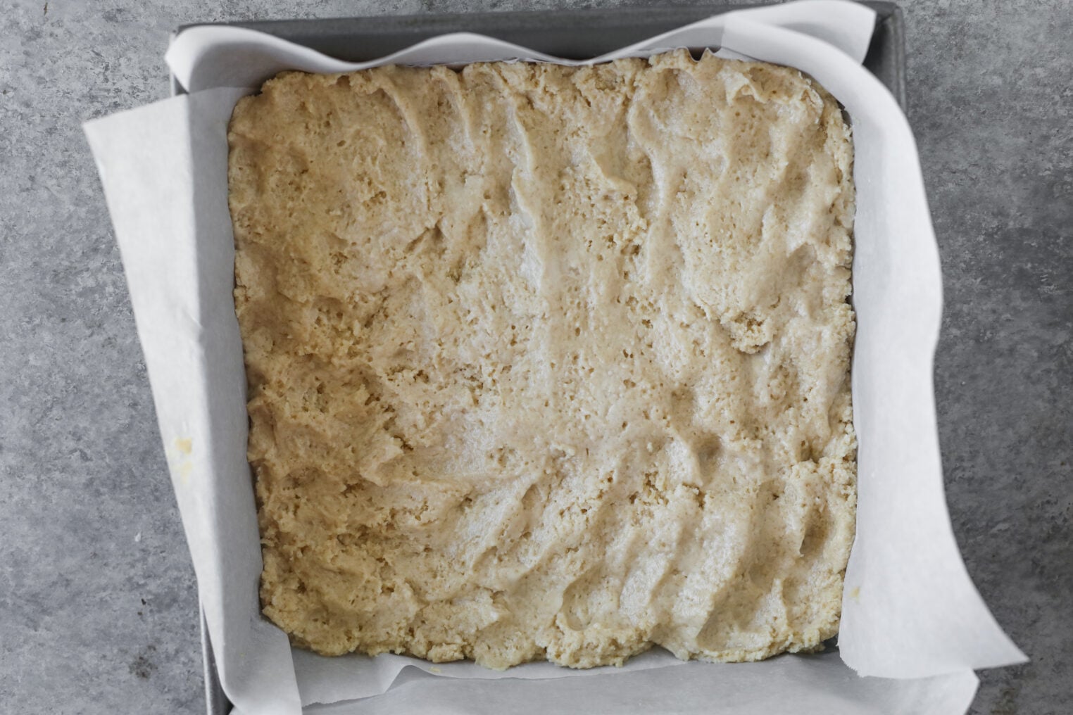 Unbaked Strawberry Coffee Cake Dough Spread Evenly In A Parchment-Lined Square Baking Pan, With A Rough Textured Surface, On A Gray Countertop.