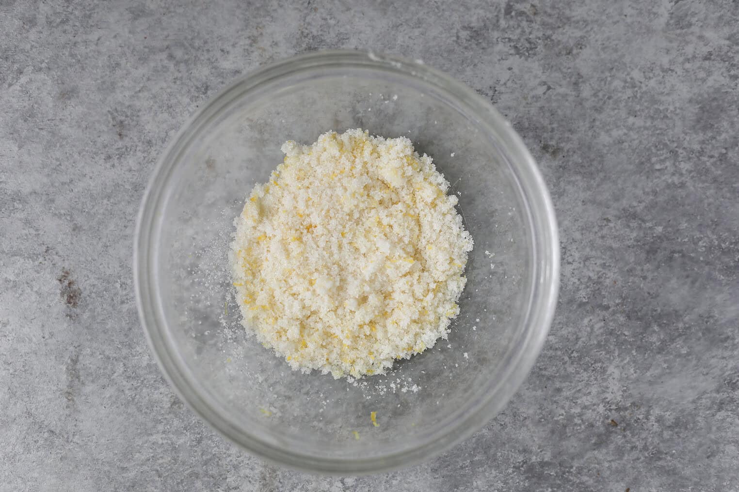 Vegan Strawberry Lemonade Scones 7 A Glass Bowl Containing A Mixture Of Finely Grated Yellow And White Ingredients, Likely Lemon Zest And Sugar For Lemonade Scones, Sits On A Gray Textured Surface.