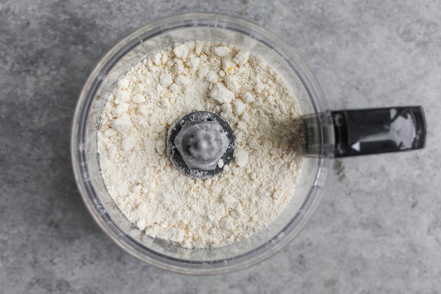 Vegan Strawberry Lemonade Scones 8 A Top-Down View Of A Food Processor Containing A Coarse, Pale Flour Mixture For Vegan Scones On A Gray Countertop.