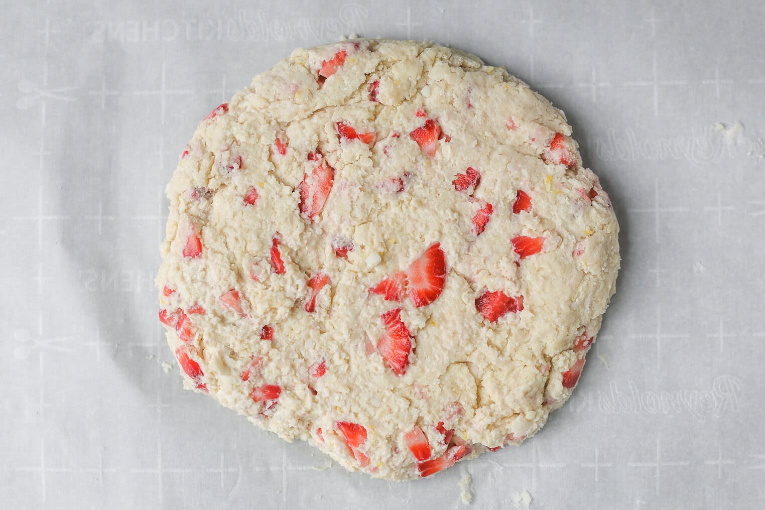 Vegan Strawberry Lemonade Scones 11 A Round, Unbaked Dough With Visible Pieces Of Fresh Strawberries—Perfect For Lemonade Scones—Is Placed On A Sheet Of Parchment Paper, Ready For Baking.