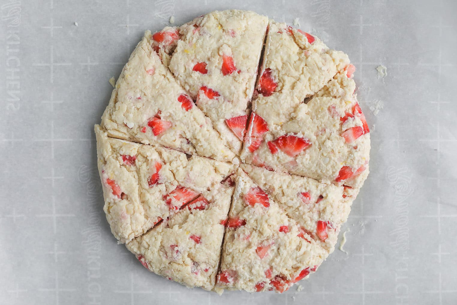 Vegan Strawberry Lemonade Scones 12 A Round Disk Of Raw Vegan Scone Dough With Visible Strawberry Pieces Is Cut Into Eight Triangular Wedges, Resting On A Sheet Of Parchment Paper.