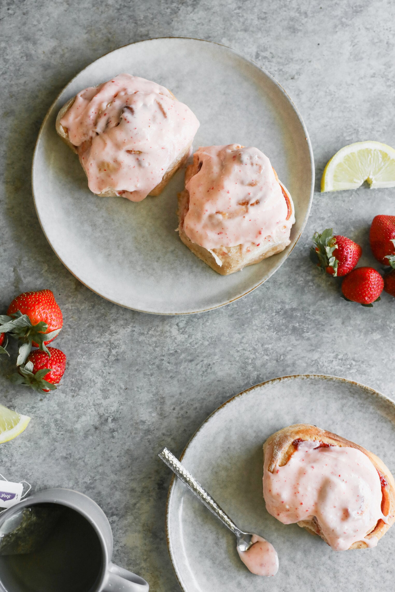 Two Plates With Fluffy Strawberry Rolls Topped With Pink Strawberry Glaze. Fresh Strawberries And Lemon Wedges Are On The Table, Along With A Mug Of Coffee And A Spoon With Extra Glaze On A Plate.