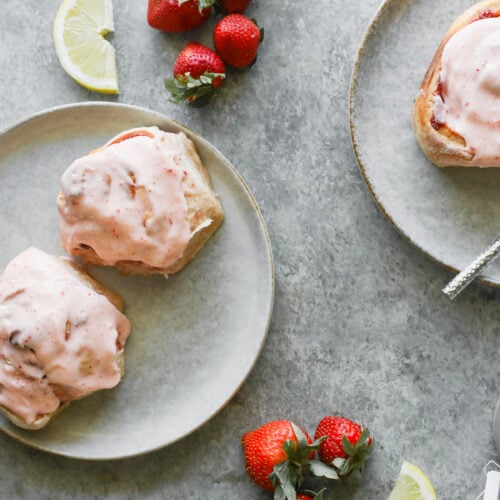 Two Plates With Strawberry Rolls Topped With Pink Strawberry Frosting; Fresh Strawberries And Lemon Slices Are Scattered Around On A Gray Surface, Along With A Spoon And A Gray Mug In The Corner.