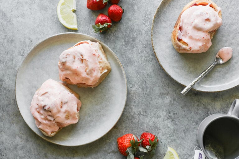 Two Plates With Strawberry Rolls Topped With Pink Strawberry Frosting; Fresh Strawberries And Lemon Slices Are Scattered Around On A Gray Surface, Along With A Spoon And A Gray Mug In The Corner.