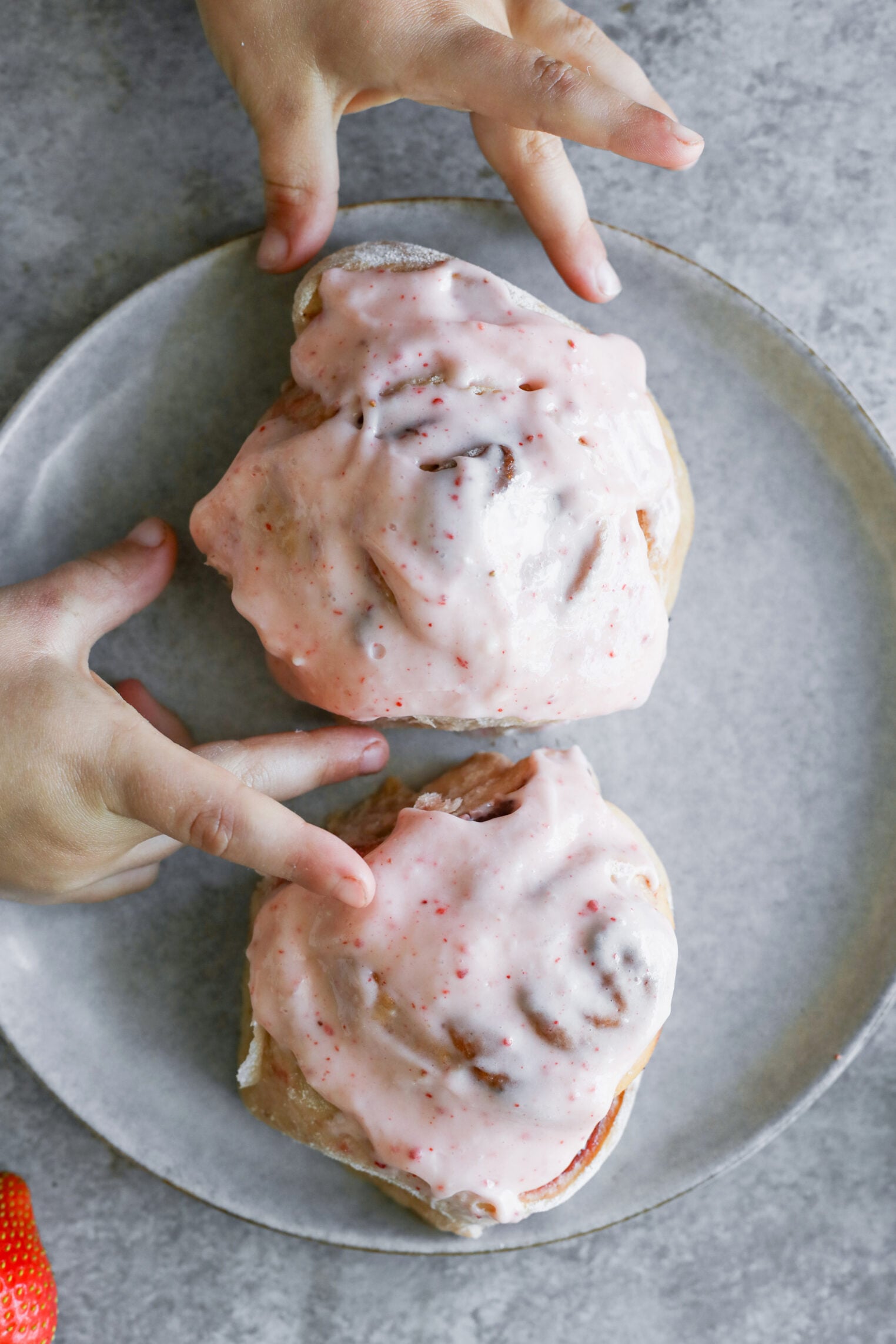 Two Small Hands Reach For Two Strawberry Rolls Topped With Pink Icing On A Gray Plate. The Background Features A Gray Surface, And Part Of A Fresh Strawberry Is Visible In The Corner.