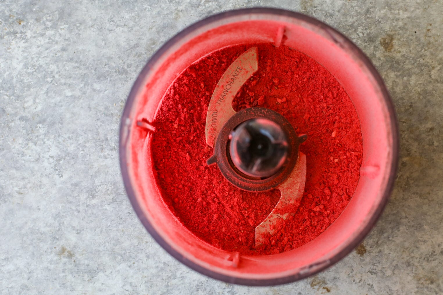 A Top-Down View Of A Grinder Filled With Bright Red Powder, Resembling The Vibrant Hue Of Strawberry Rolls, Showcases Metal Blades And Evenly Distributed Powder Against A Gray Textured Background.