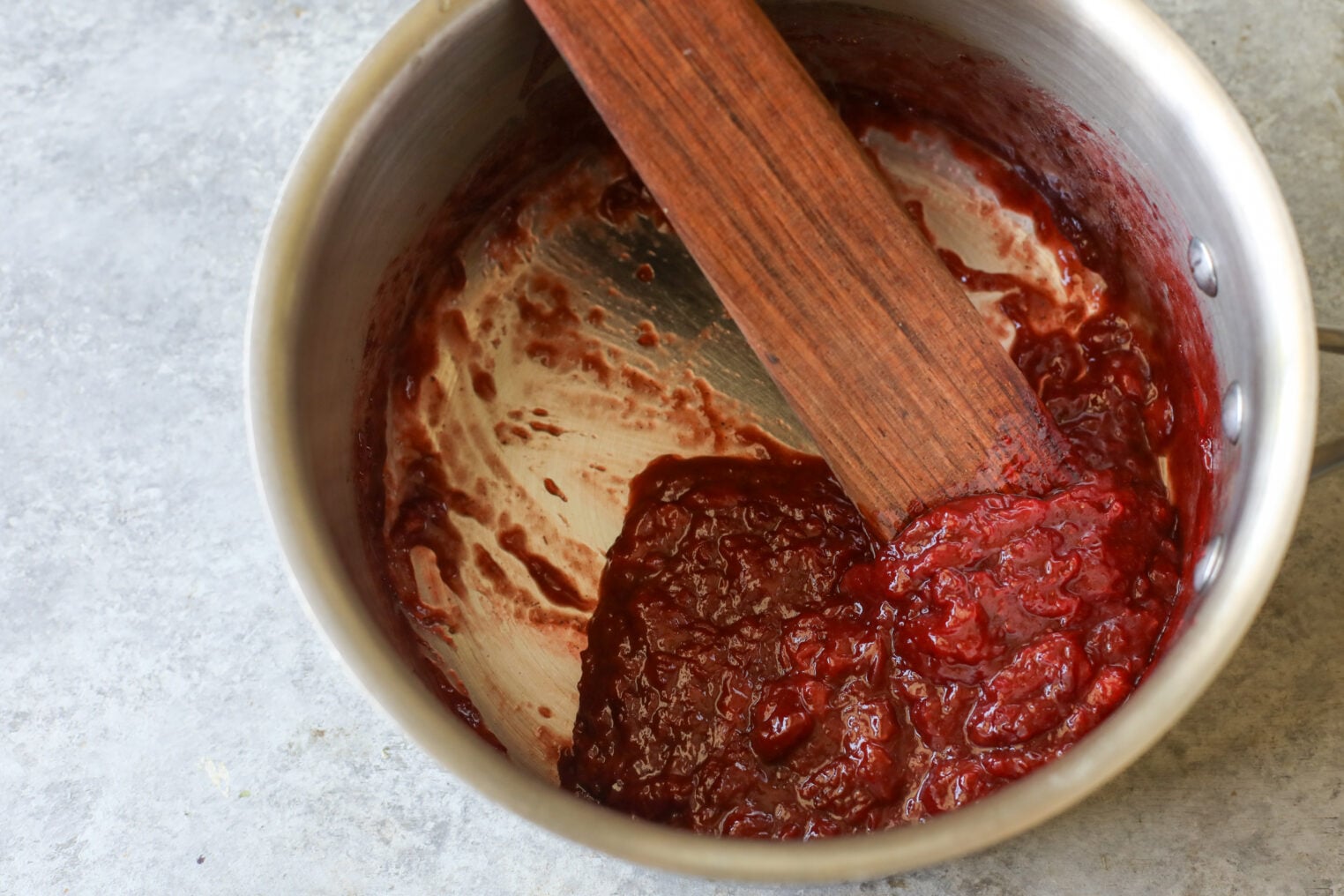 A Saucepan Containing Thick Red Fruit Jam Or Sauce, Perfect For Making Strawberry Rolls, Is Partially Scraped With A Wooden Spatula And Sits On A Light Gray Surface.