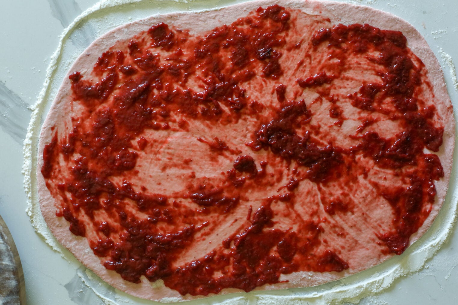 Oval-Shaped Dough Rolled Out On A Floured Surface, Spread With A Sweet Layer Of Red Fruit Filling—Perfect For Making Delicious Strawberry Rolls, Ready For Further Preparation Or Baking.