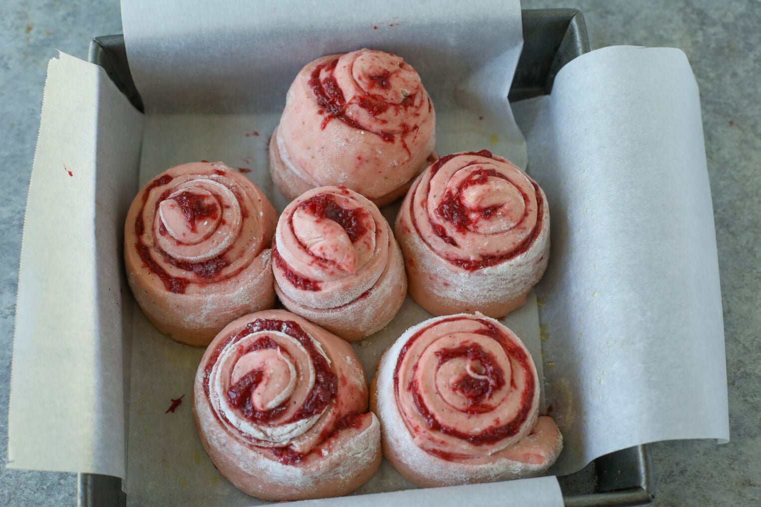 Unbaked Pink Raspberry And Strawberry Rolls Are Arranged In A Parchment-Lined Square Baking Pan On A Gray Surface, Ready To Be Baked. The Dough Is Lightly Dusted With Flour And Swirled With Vibrant Berry Filling.