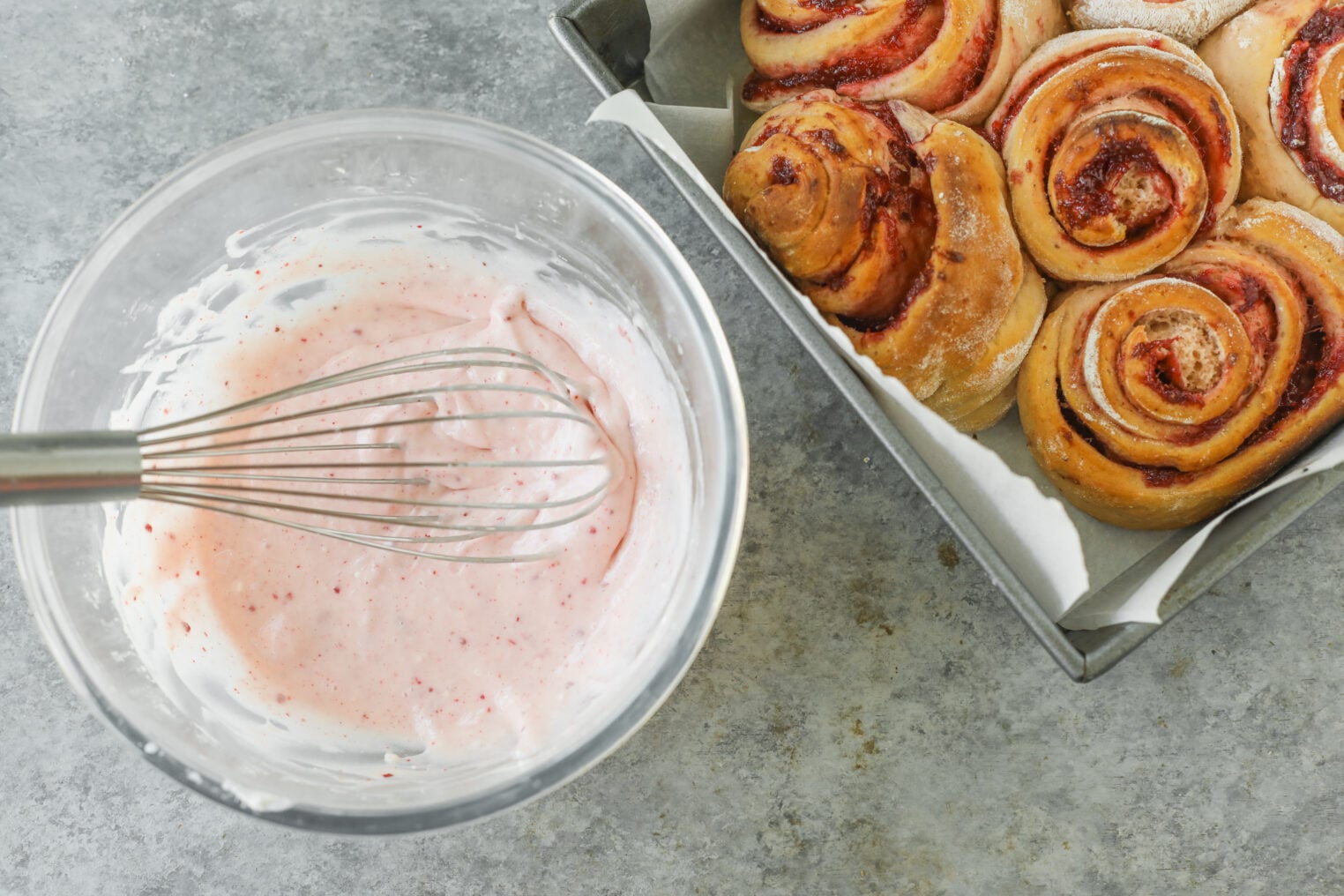 A Glass Bowl With Pink Icing And A Whisk Sits Next To A Baking Pan Filled With Freshly Baked Strawberry Rolls On A Gray Countertop, Ready To Be Glazed For The Perfect Strawberry Rolls Treat.