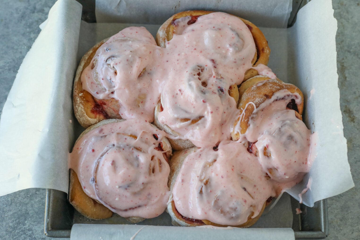A Square Baking Pan Lined With Parchment Paper Holds Six Strawberry Rolls Generously Covered In Pink Icing. The Rolls Are Slightly Touching, And The Creamy Strawberry Icing Is Spread Unevenly For A Homemade Look.