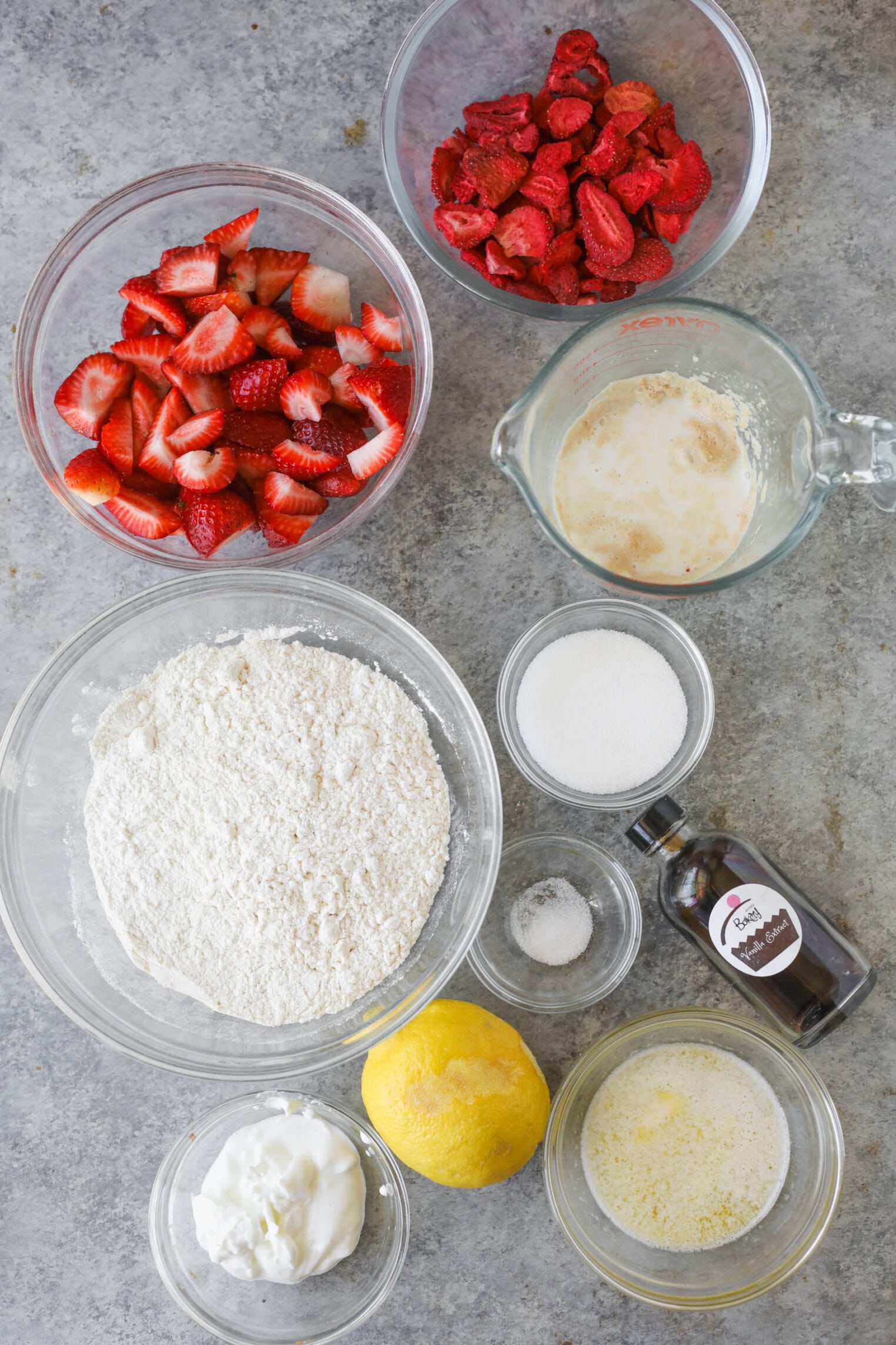 Bowls Containing Ingredients For Strawberry Rolls—Sliced Strawberries, Freeze-Dried Strawberries, Flour, Sugar, Salt, Baking Powder, A Lemon, Melted Butter, Yogurt, Milk With Lemon Juice, And Vanilla Extract—Sit On A Gray Countertop.