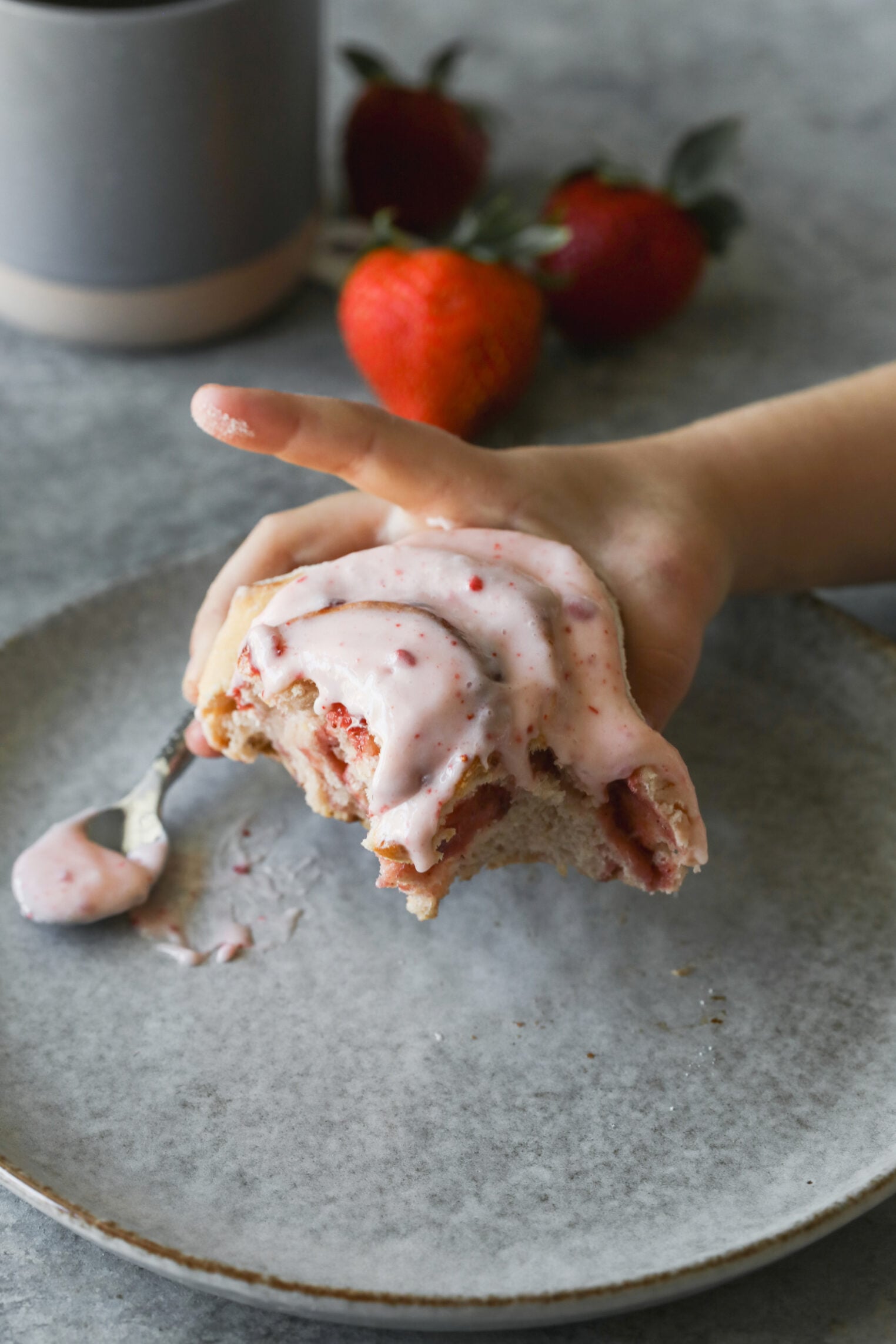 A Hand Holds A Partially Eaten Strawberry Roll Topped With Pink Strawberry Icing Over A Gray Plate, With A Spoon Of Icing And Fresh Strawberries In The Background.