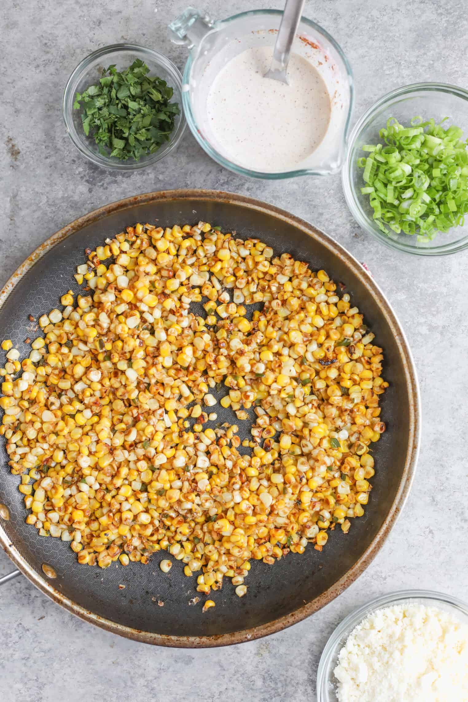 Easy Street Corn Tostadas 2 A Skillet With Sautéed Street Corn Sits On A Gray Surface, Surrounded By Bowls Of Chopped Cilantro, Sliced Green Onions, Crumbled Cheese, And A Measuring Cup Of Creamy Sauce—Perfect Toppings For Tostadas.