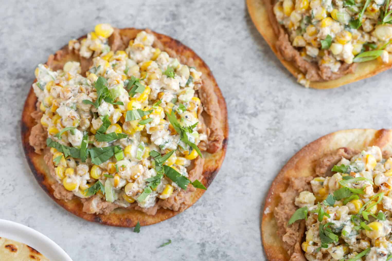 Easy Street Corn Tostadas 8 A Close-Up Of Tostadas Topped With Refried Beans, Street Corn Salad, Crumbled Cheese, And Chopped Cilantro On A Light Gray Surface.
