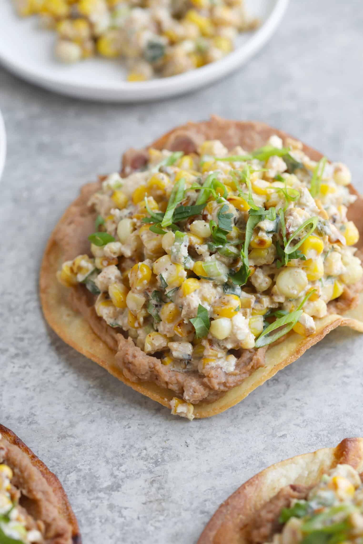 Easy Street Corn Tostadas 9 A Close-Up Of Street Corn Tostadas Topped With Refried Beans, A Creamy Corn Mixture, And Garnished With Sliced Green Onions, Served On A Gray Surface.