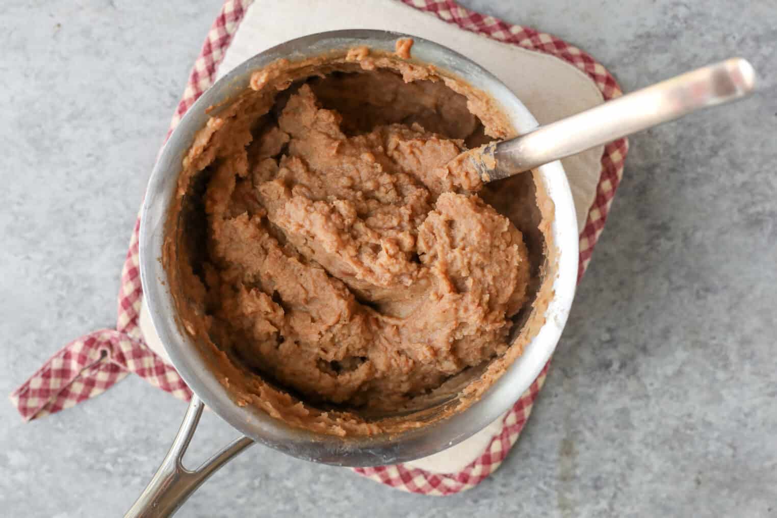Easy Street Corn Tostadas 6 A Pot Of Creamy Refried Beans With A Spoon Rests On A Red And White Checkered Cloth, Ready To Be Spread Onto Crisp Tostadas For The Perfect Street Corn Tostadas Experience.