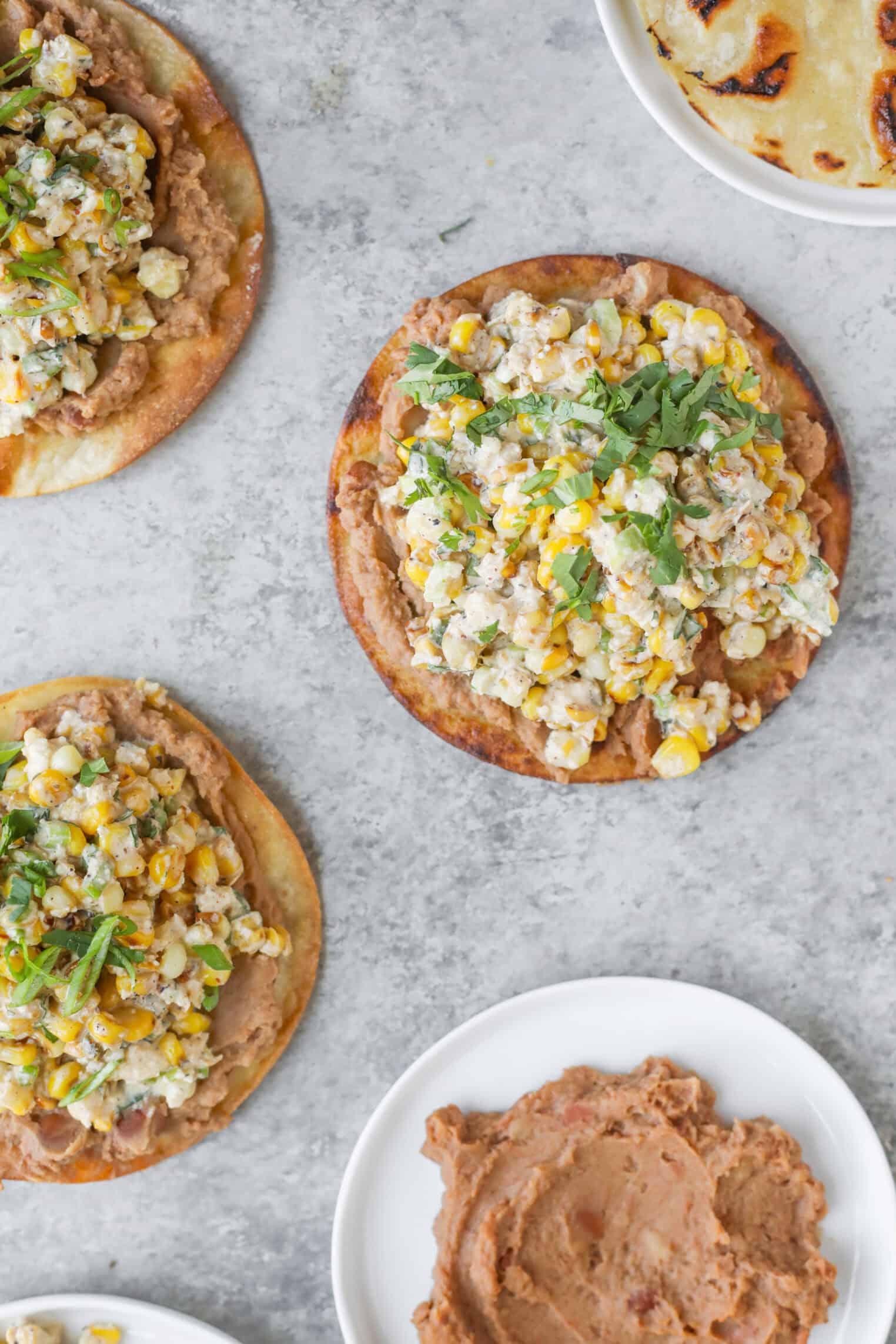 Easy Street Corn Tostadas 3 Three Street Corn Tostadas Topped With Refried Beans And A Corn And Herb Mixture Are Arranged On A Light Gray Surface, With An Extra Bowl Of Refried Beans Nearby. One Tostada Is Garnished With Fresh Herbs.