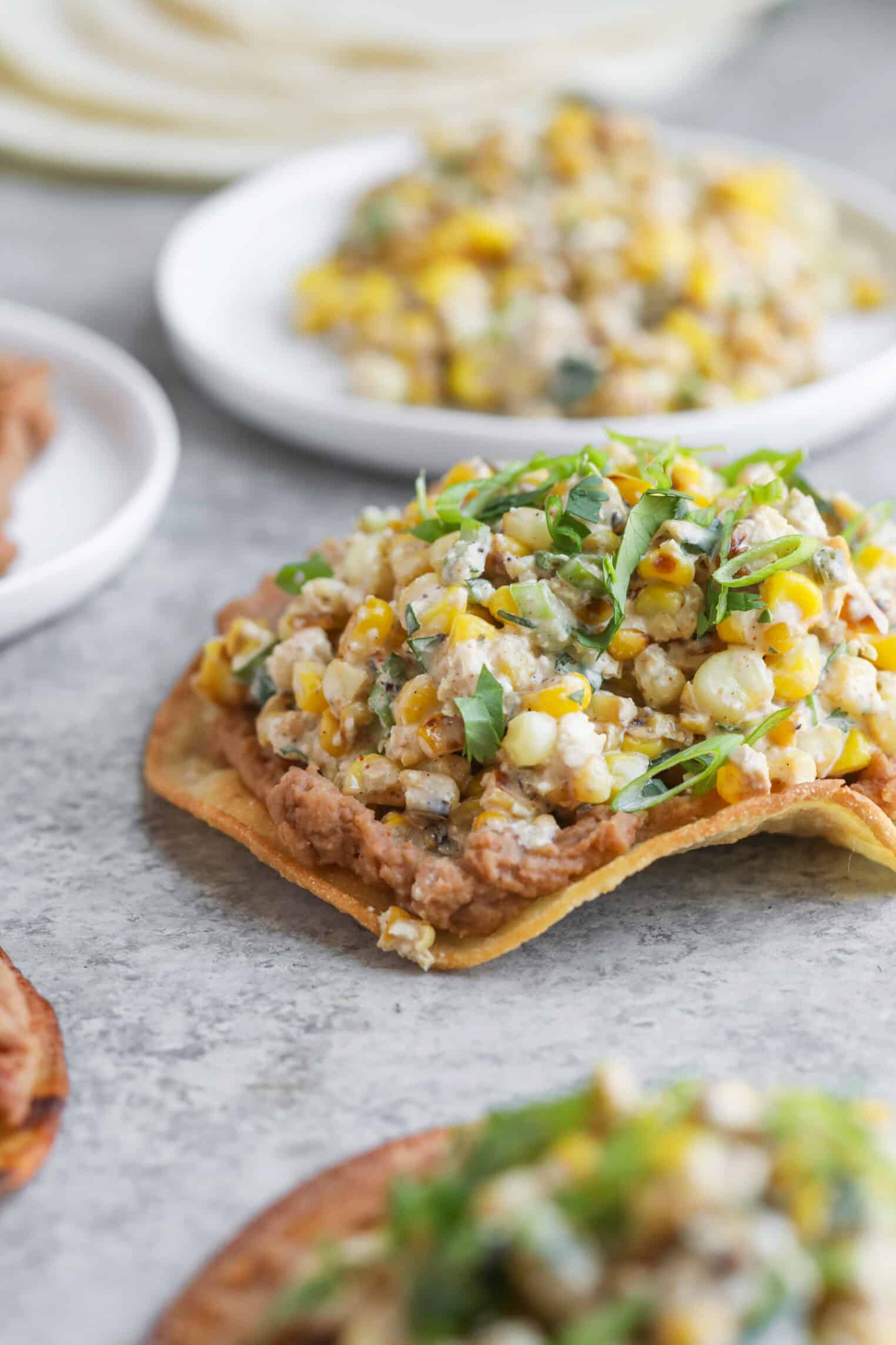 Easy Street Corn Tostadas 1 A Close-Up Of A Tostada Topped With Refried Beans, Street Corn Salad, And Fresh Herbs, Set On A Gray Surface With Other Delicious Tostadas Blurred In The Background.