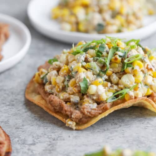 Easy Street Corn Tostadas 11 A Close-Up Of A Tostada Topped With Refried Beans, Street Corn Salad, And Fresh Herbs, Set On A Gray Surface With Other Delicious Tostadas Blurred In The Background.