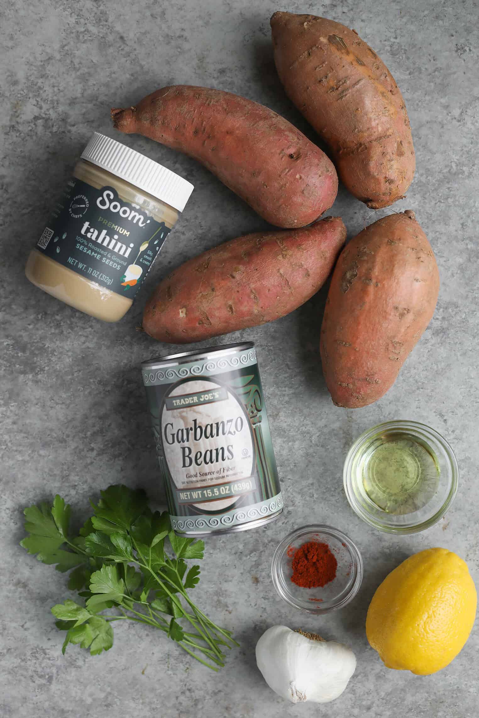 Vegan Stuffed Sweet Potatoes 3 Ingredients For Vegan Stuffed Sweet Potatoes Are Arranged On A Gray Surface: Sweet Potatoes, A Jar Of Tahini, Garbanzo Beans, Parsley, A Small Bowl Of Oil, A Lemon, Garlic, And A Small Bowl Of Paprika.