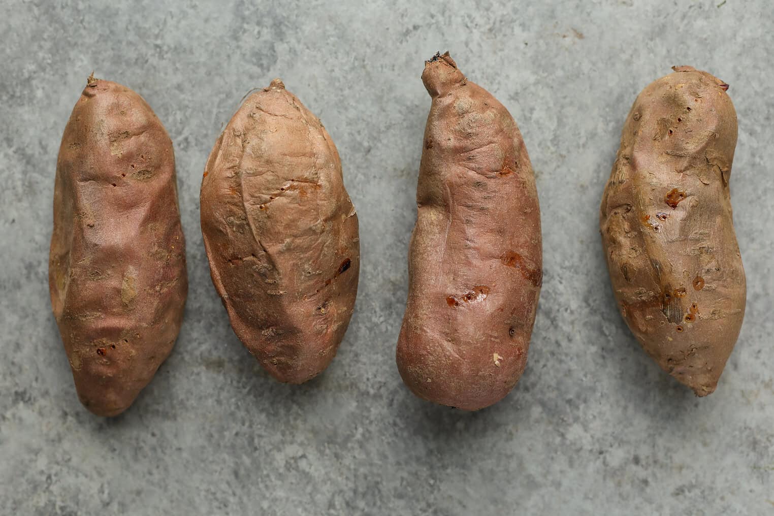 Vegan Stuffed Sweet Potatoes 5 Four Whole, Unpeeled Sweet Potatoes With Rough, Brownish-Orange Skin Are Arranged In A Row On A Gray, Textured Surface—Perfect For Making Vegan Stuffed Sweet Potatoes.
