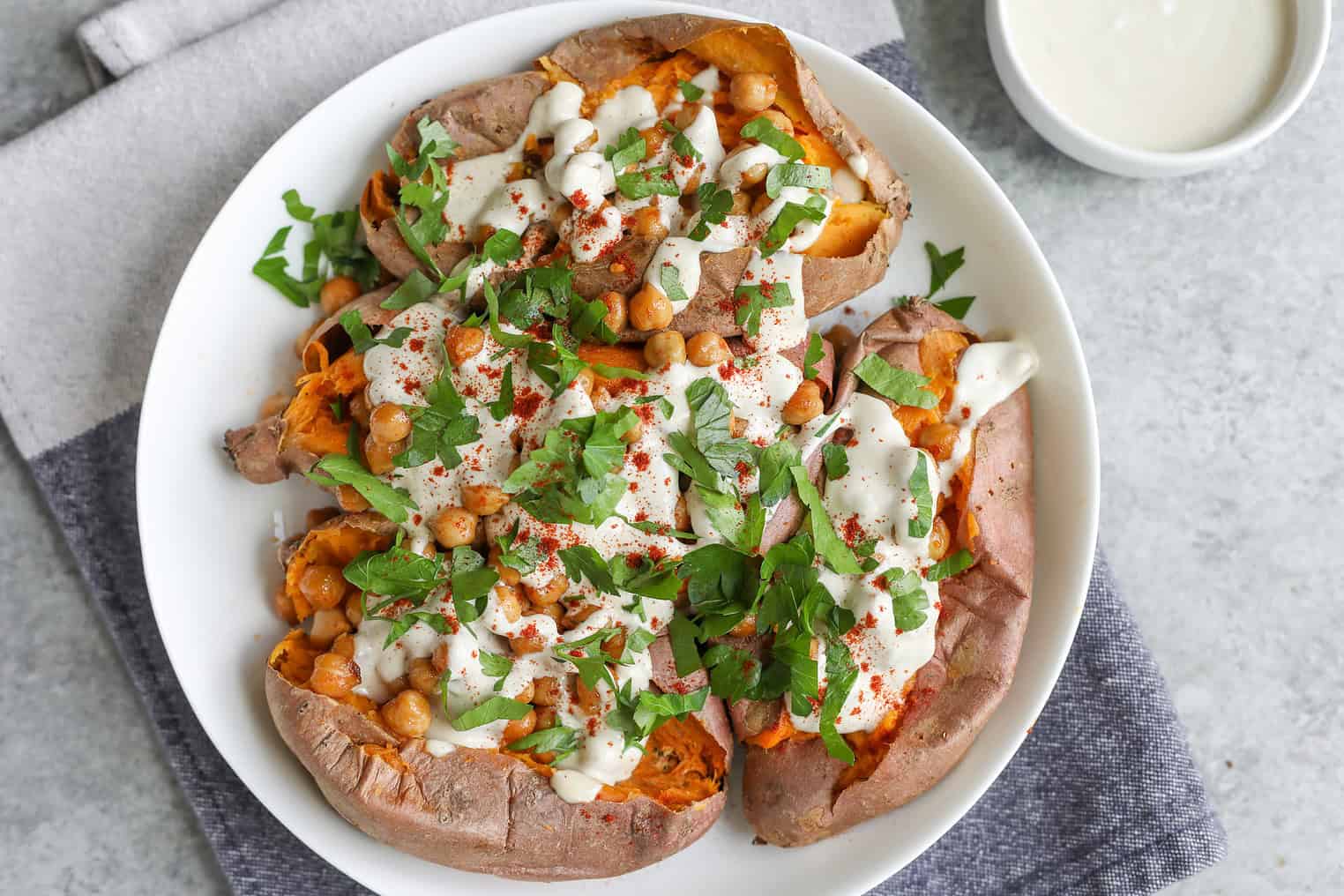 Vegan Stuffed Sweet Potatoes 9 A Plate Of Vegan Stuffed Sweet Potatoes Topped With Chickpeas, Chopped Fresh Parsley, Creamy White Sauce, And A Sprinkle Of Paprika, Served On A Gray Napkin With A Small Bowl Of Extra Sauce Nearby.