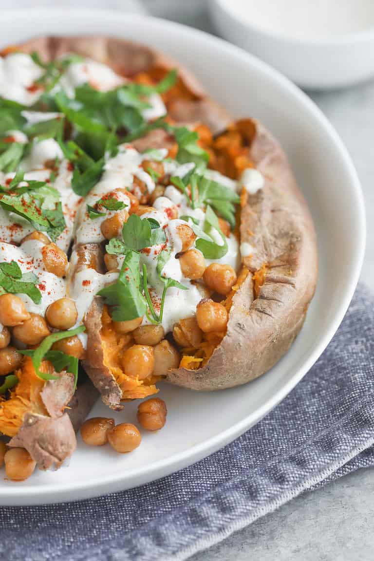 A Baked Sweet Potato Split Open And Filled With Chickpeas, Drizzled With A Creamy White Sauce, And Topped With Fresh Herbs—These Vegan Stuffed Sweet Potatoes Are Served On A White Plate With A Gray Textured Napkin Underneath.
