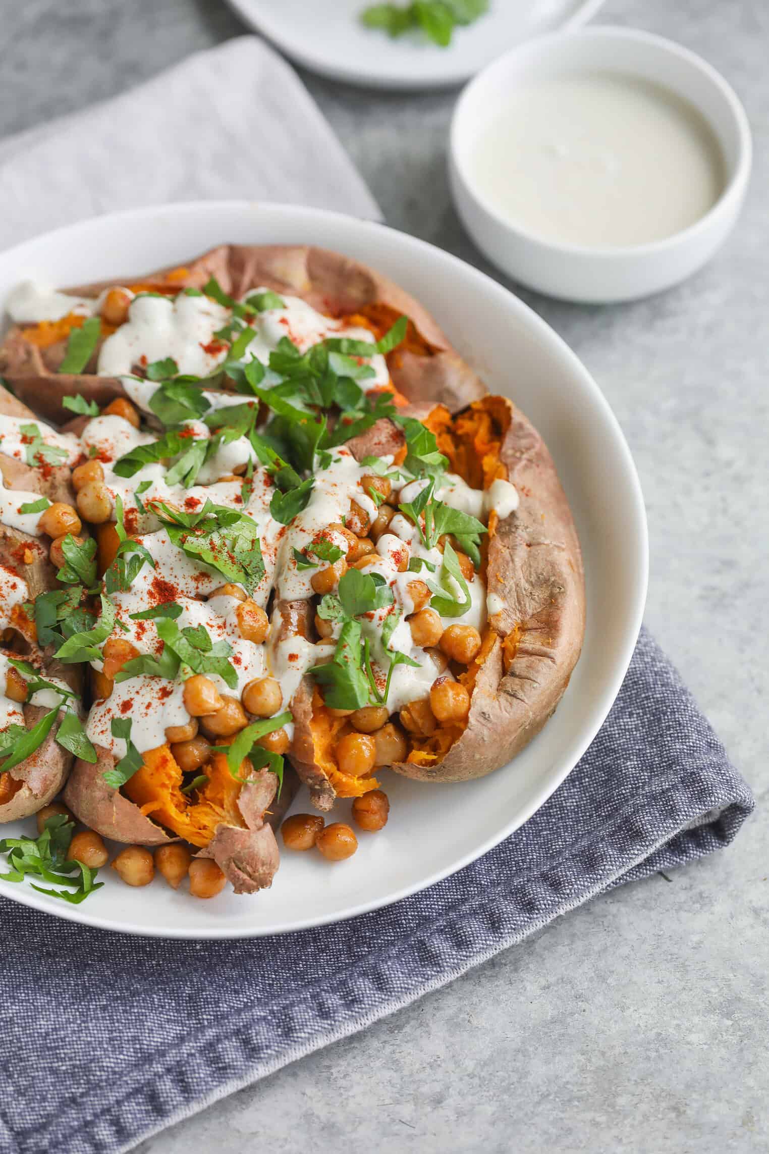 Vegan Stuffed Sweet Potatoes 10 A Baked Stuffed Sweet Potato Split Open And Topped With Chickpeas, Chopped Fresh Herbs, And Creamy White Sauce, Served On A White Plate With A Gray Napkin. A Small Bowl Of Extra Sauce Is In The Background.