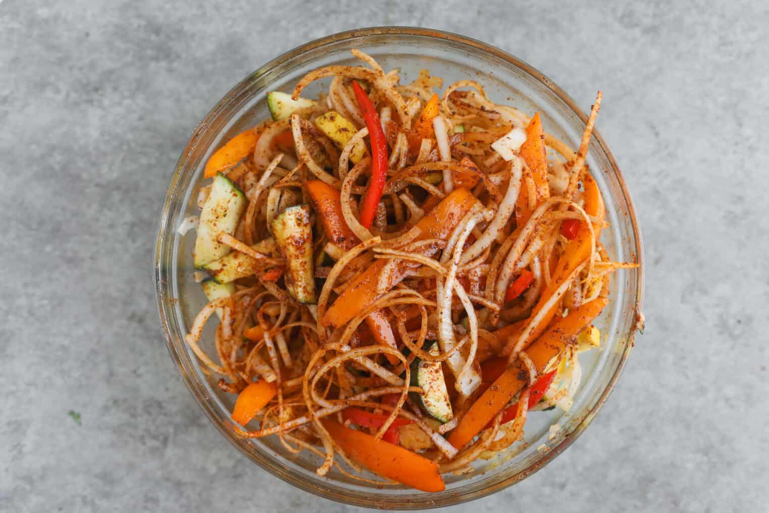 A Glass Bowl Filled With A Colorful Salad Of Thinly Sliced Vegetables—Perfect For Summer Fajita Bowls. Orange And Red Bell Peppers, Onions, And Zucchini Are Coated In Spices And Served On A Gray Surface.