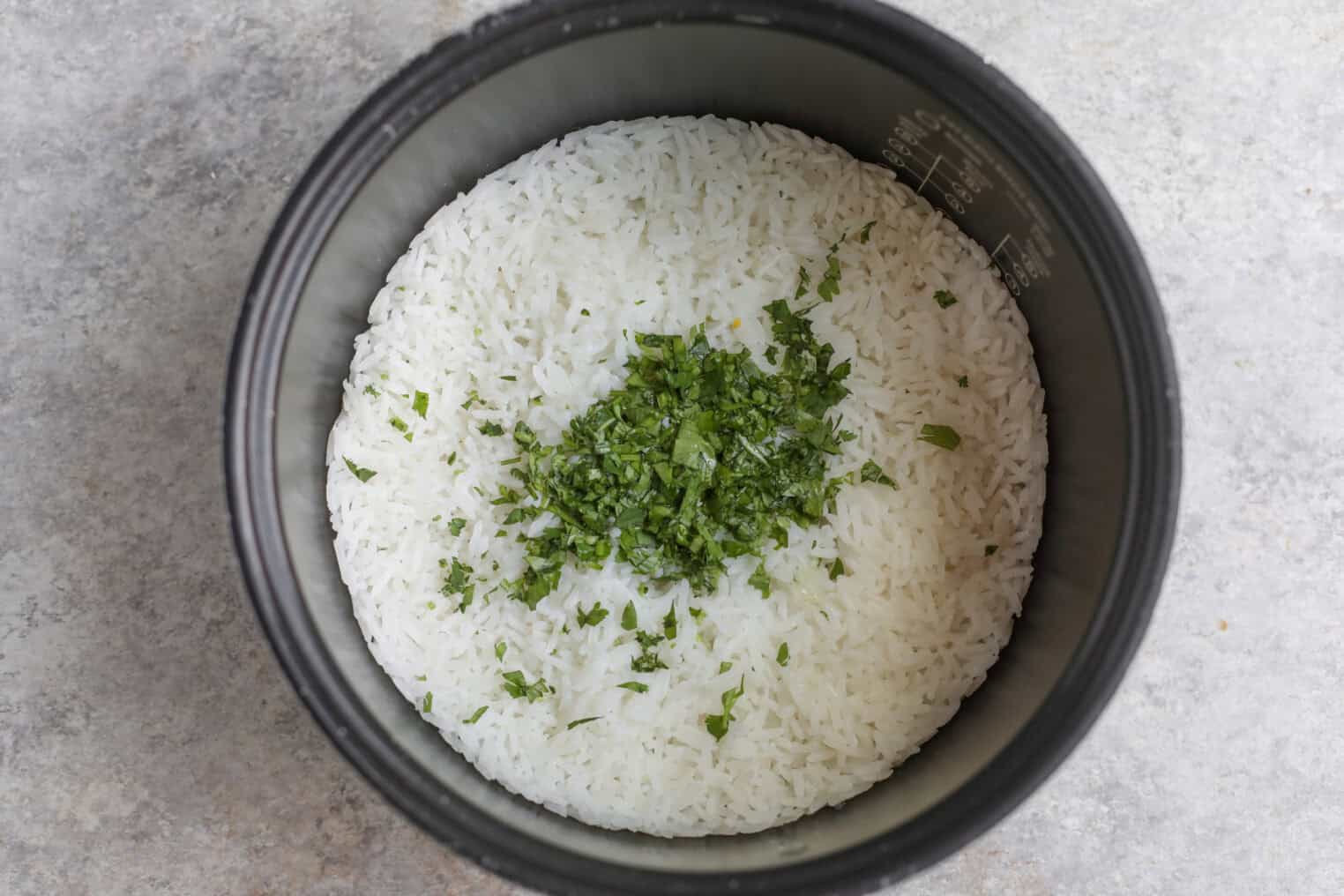 A Rice Cooker Filled With Cooked White Rice, Topped With A Small Pile Of Chopped Fresh Herbs, Perfect As A Base For Summer Fajita Bowls, Sits On A Gray Countertop.