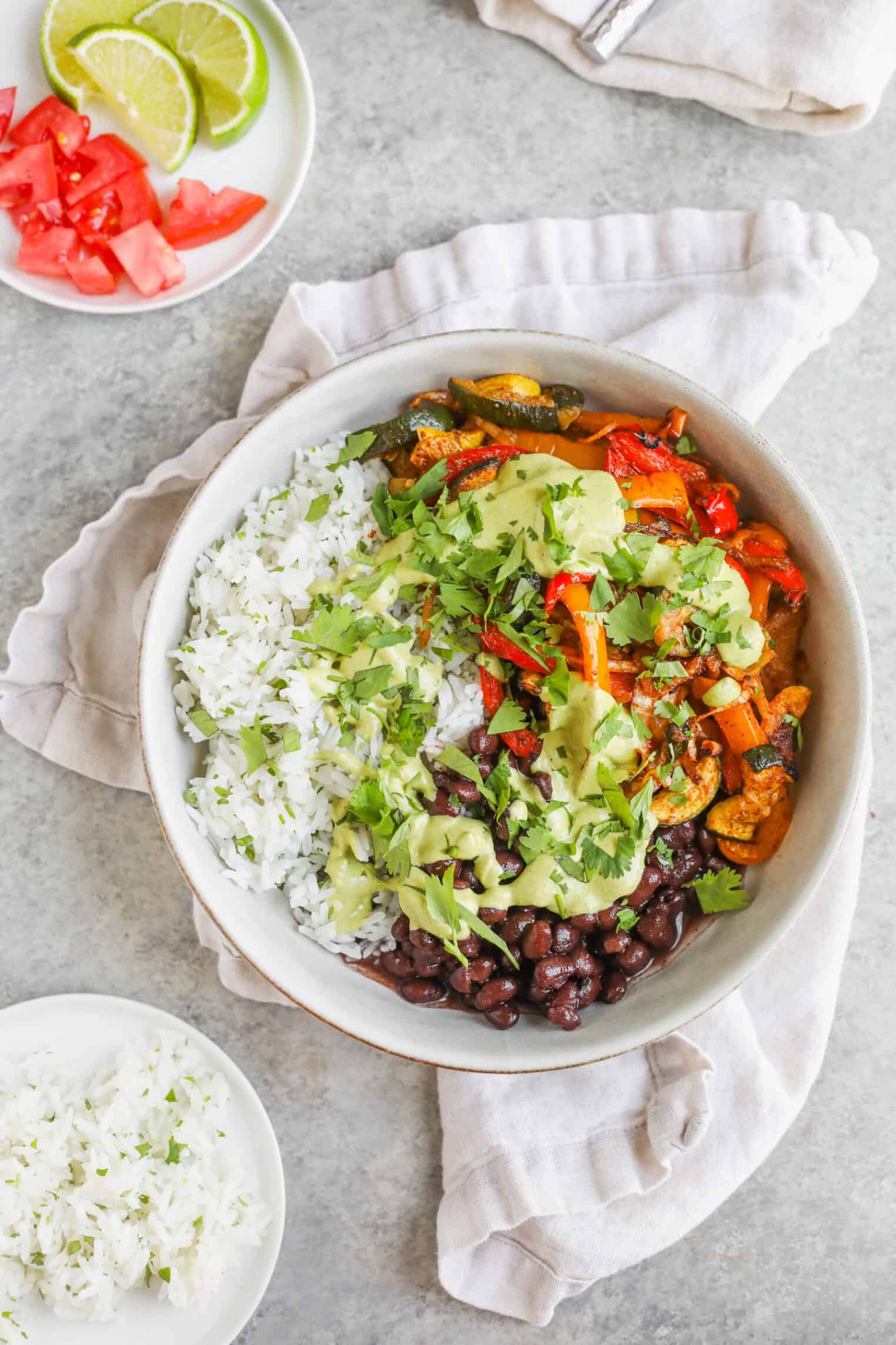 A Bowl Filled With White Rice, Black Beans, Roasted Vegetables, And Topped With Fresh Cilantro And Creamy Green Sauce Makes These Summer Fajita Bowls Shine. Nearby Are Small Dishes With Lime Wedges, Chopped Tomatoes, And Extra Rice.