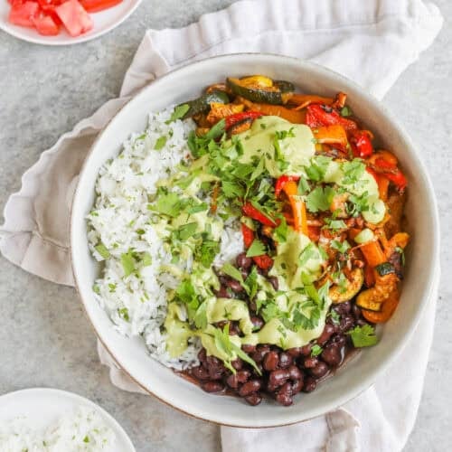 A Bowl With White Rice, Black Beans, Sautéed Vegetables, And Creamy Sauce, Topped With Fresh Cilantro—These Vegetable Fajita Bowls Are Served With Plates Of Sliced Lime, Diced Tomato, And Extra Rice On A Light Napkin And Countertop.