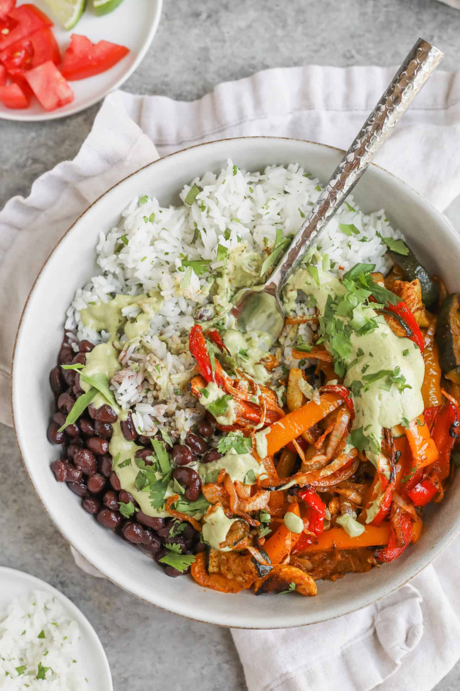 A Bowl Filled With White Rice, Black Beans, Sautéed Bell Peppers And Onions, Topped With Creamy Green Sauce And Fresh Cilantro—A Perfect Summer Fajita Bowl. A Spoon Rests Inside, With Plates Of Tomatoes And Lime Slices Nearby.