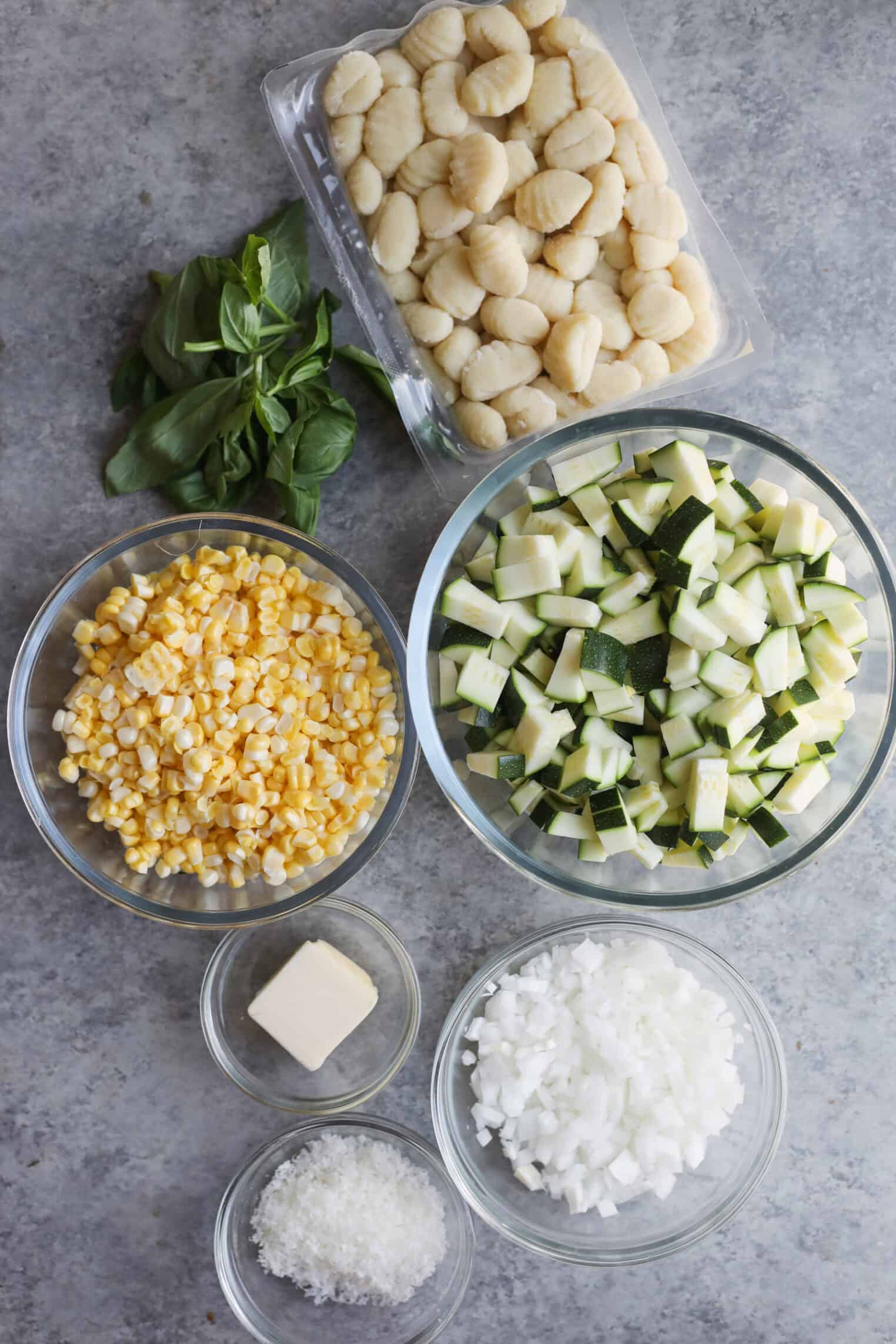 An Overhead View Of Ingredients For Summer Gnocchi On A Gray Surface: Fresh Gnocchi, Basil Leaves, Diced Zucchini For Zucchini Sauce, Fresh Corn Kernels, Diced Onion, A Pat Of Butter, And Grated Cheese In Separate Bowls.