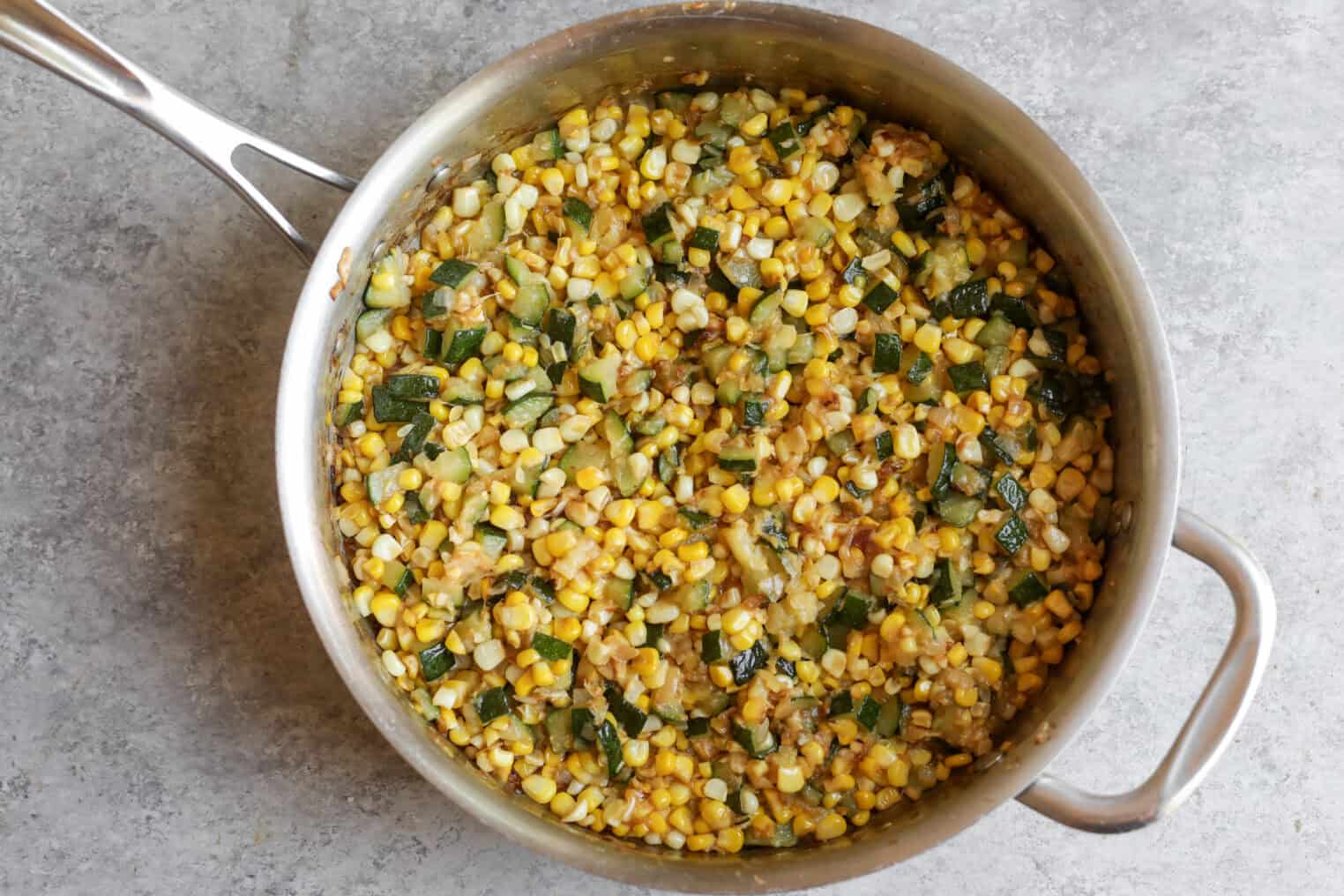 A Stainless Steel Pan Filled With Sautéed Fresh Corn, Diced Zucchini, And Onions Sits On A Gray Countertop. The Vegetables Are Mixed Together And Appear Cooked And Slightly Browned.
